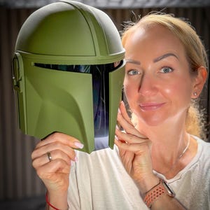 May include: An olive green Mandalorian helmet with a visor. The helmet has a rounded top and angular features, with a dark interior visible through the visor. The helmet is held up in front of a person.