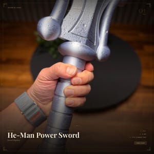 Pode incluir: Uma réplica prateada da espada He-Man Power Sword, segurada por uma mão. A espada tem um design detalhado de punho e lâmina. O texto "He-Man Power Sword" e "Sword Replica" é visível na parte inferior da imagem.