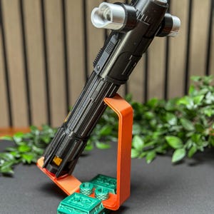 Op de afbeelding: Een zwart en zilver lichtzwaard rekwisiet wordt getoond op een oranje standaard met groene decoratieve stukken. Het lichtzwaard heeft een gedetailleerd ontwerp en is omhoog gekanteld. De standaard en decoratieve stukken staan op een donkere ondergrond met groen op de achtergrond.