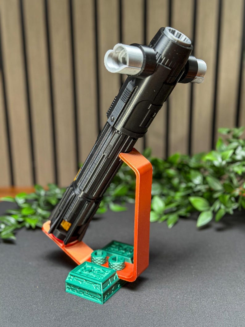 Op de afbeelding: Een zwart en zilver lichtzwaard rekwisiet wordt getoond op een oranje standaard met groene decoratieve stukken. Het lichtzwaard heeft een gedetailleerd ontwerp en is omhoog gekanteld. De standaard en decoratieve stukken staan op een donkere ondergrond met groen op de achtergrond.