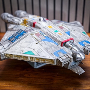Puede incluir: Modelo detallado de una nave espacial, principalmente plateada con detalles azules, rojos, amarillos y verdes. El modelo presenta una cabina y un diseño de motor detallados, expuesto sobre una superficie de madera.