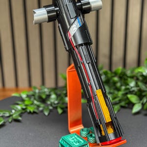 Op de afbeelding: Een zwart en zilver lichtzwaard met een kruisbeschermingsontwerp, met rode en blauwe interne bedradingsdetails. Het lichtzwaard rust op een oranje standaard met een groene decoratieve basis, met een gedetailleerde, futuristische esthetiek.