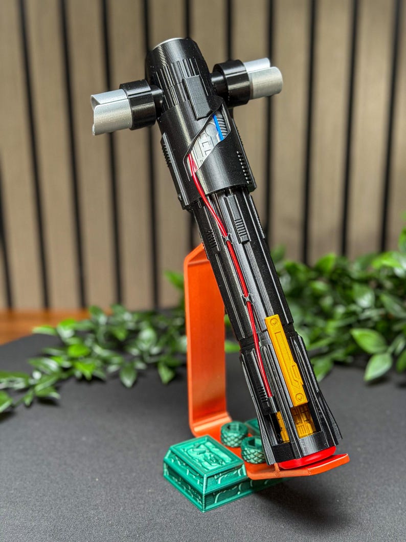 Op de afbeelding: Een zwart en zilver lichtzwaard met een kruisbeschermingsontwerp, met rode en blauwe interne bedradingsdetails. Het lichtzwaard rust op een oranje standaard met een groene decoratieve basis, met een gedetailleerde, futuristische esthetiek.
