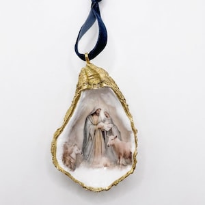 May include: A decorative oyster shell ornament featuring a painted nativity scene with figures in robes and animals. The shell has a metallic gold painted edge and is suspended by a dark blue velvet ribbon tied in a loop at the top for hanging.