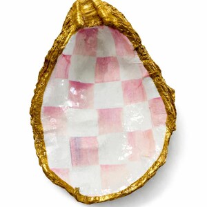 May include: Decorative oyster shell with a gold-painted rim. The interior features a pink and white checkered pattern. The shell is a unique decorative item, perfect for holding small items or as a coastal-themed accent.