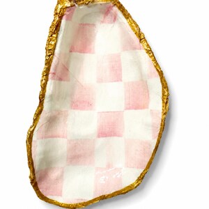 May include: A decorative oyster shell featuring a hand-painted pink and white checkered pattern on the interior surface. The outer edge of the shell is finished with a textured, metallic gold painted rim, highlighting the natural organic shape.