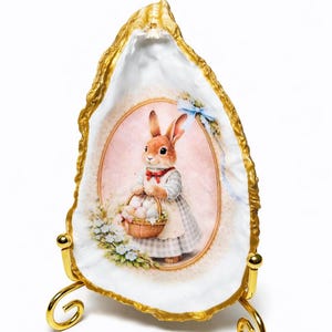 May include: An Easter-themed decorative oyster shell with a gold-painted edge, supported by a gold stand. The shell features a painted image of a bunny holding a basket of eggs, with a pink background and floral accents.