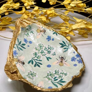 May include: A decorative oyster shell with a gold-painted rim. The interior features a light green fabric with a floral and bee pattern. The design includes blue flowers, white blossoms, and detailed bee illustrations.