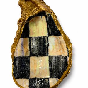 May include: An oyster shell with a metallic gold painted exterior and a hand-painted interior featuring a black and white checkered pattern. The surface of the interior shows subtle brushstrokes and hints of light pink and yellow tones.