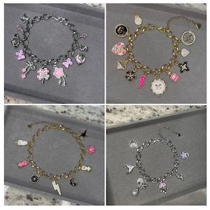 May include: Four charm bracelets with various charms. The bracelets are silver and gold-toned, with charms in pink, black, white, and other colors. Charms include stars, flowers, hearts, and other decorative shapes. The bracelets are displayed on a gray surface.