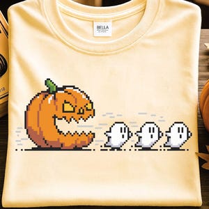 May include: A light yellow t-shirt featuring a pixelated Halloween design. The graphic shows an orange pumpkin chasing three white ghosts. The shirt has a crew neck and a Bella label.