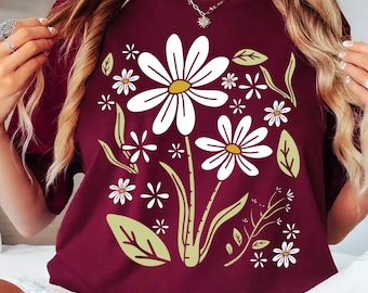 May include: Maroon t-shirt featuring a white daisy floral design with yellow centers and green leaves. The shirt is a crew neck style. The word "Maroon" is in a circle in the lower left corner.
