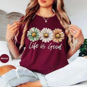 May include: Maroon t-shirt featuring three daisy flowers in green, white, and orange with yellow centers. The words "Life is Good" are written in white script below the flowers. The shirt is a crew neck style.
