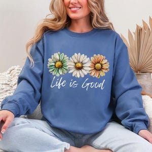 May include: A blue sweatshirt with the phrase "Life is Good" in white script. The sweatshirt features a daisy design with green, white, and orange petals. The daisies are arranged horizontally across the chest.