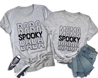 May include: Two gray t-shirts with black text that reads "DADA SPOOKY DADA" and "MAMA SPOOKY MAMA". The shirt on the left has black sneakers. The shirt on the right has blue jeans.