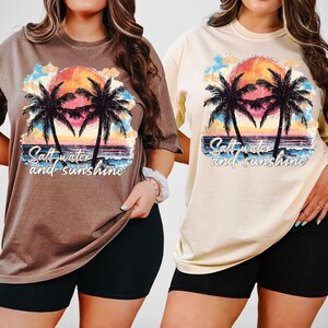 Puede incluir: Dos camisetas, una marr&oacute;n y otra crema, con un dise&ntilde;o de puesta de sol tropical. El gr&aacute;fico muestra palmeras, una colorida puesta de sol, olas del oc&eacute;ano y el texto "Salt water and sunshine".