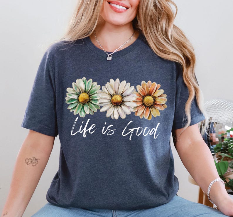 May include: A navy blue t-shirt featuring three daisy flowers in green, white, and orange with yellow centers. The words "Life is Good" are written in a cursive font below the flowers. The shirt has short sleeves.
