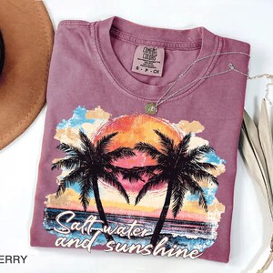 Puede incluir: Camiseta de color bayas con un gr&aacute;fico de puesta de sol tropical con palmeras y el texto "Saltwater and Sunshine". La camiseta tiene cuello redondo y una etiqueta peque&ntilde;a. Un collar plateado con un colgante est&aacute; sobre la camiseta.