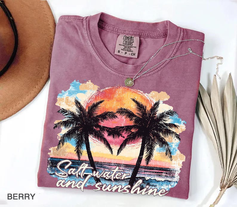 Puede incluir: Camiseta de color bayas con un gr&aacute;fico de puesta de sol tropical con palmeras y el texto "Saltwater and Sunshine". La camiseta tiene cuello redondo y una etiqueta peque&ntilde;a. Un collar plateado con un colgante est&aacute; sobre la camiseta.