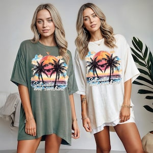Puede incluir: Dos camisetas holgadas, una verde oliva y otra blanquecina, cada una con un gr&aacute;fico de puesta de sol tropical, palmeras y el texto "Salt water and sunshine". Las camisetas tienen mangas cortas y un corte holgado.