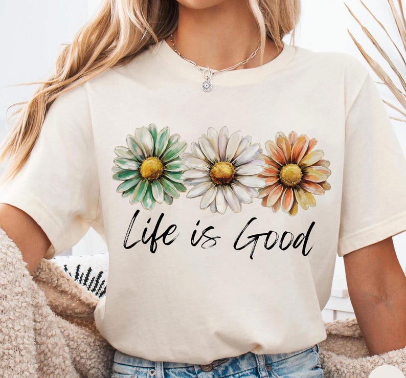 May include: Cream-colored t-shirt featuring three daisy flowers in green, white, and orange with yellow centers. The phrase "Life is Good" is written in black script below the flowers. The shirt has short sleeves.