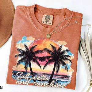 Puede incluir: Una camiseta de color coral con un dise&ntilde;o de puesta de sol tropical, palmeras y el texto "Salt water and sunshine". El gr&aacute;fico incluye una colorida puesta de sol, olas del oc&eacute;ano y un cielo azul y amarillo. Un collar dorado est&aacute; sobre la camiseta.