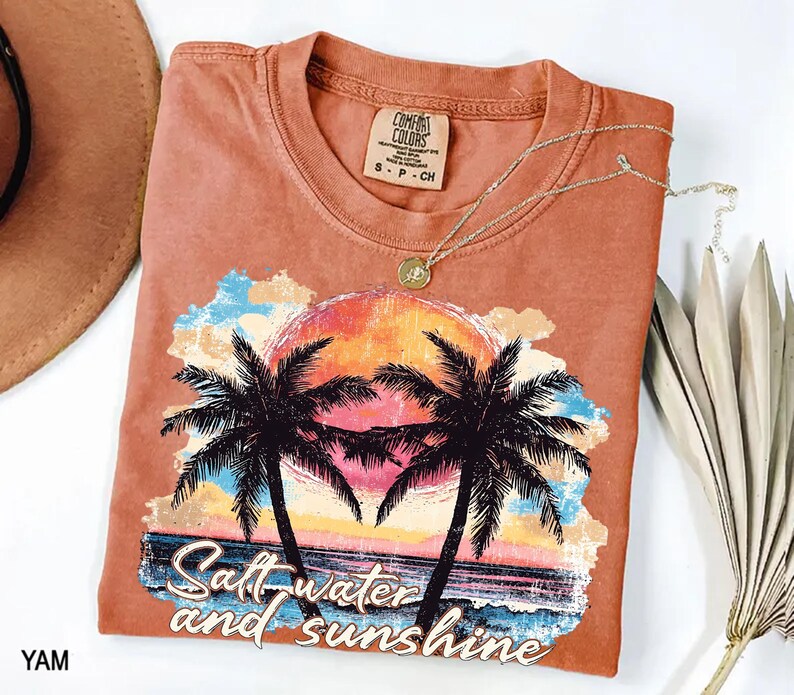 Puede incluir: Una camiseta de color coral con un dise&ntilde;o de puesta de sol tropical, palmeras y el texto "Salt water and sunshine". El gr&aacute;fico incluye una colorida puesta de sol, olas del oc&eacute;ano y un cielo azul y amarillo. Un collar dorado est&aacute; sobre la camiseta.