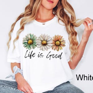 May include: White t-shirt featuring three daisy flowers in shades of green, white, and orange, with yellow centers. The phrase "Life is Good" is written in black script below the flowers. The shirt is a casual style.