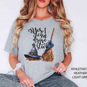 May include: Light grey t-shirt with the text "Yes, I can drive a stick!" in black script. The graphic includes a witch's hat with leaves and a broom. The shirt is heathered and made of a soft material.