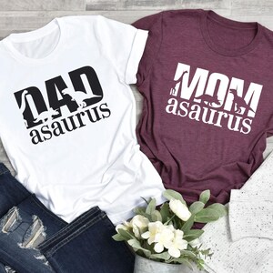 May include: Two t-shirts, one white and one burgundy, with dinosaur-themed text. The white shirt says "DAD asaurus" in black, and the burgundy shirt says "MOM asaurus" in white. Dark blue jeans and a bouquet of flowers are in the foreground.