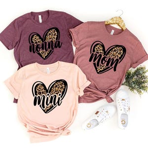 May include: Three short-sleeved t-shirts in shades of pink and burgundy. Each shirt features a heart design with a leopard print fill and a word in black script: "Nonna", "Mom", and "Mini". White sneakers are next to the shirts.
