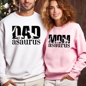 May include: A white sweatshirt with the text "DAD asaurus" in black, featuring dinosaur silhouettes. Next to it, a pink sweatshirt with "MOM asaurus" in black, also with dinosaur silhouettes. The sweatshirts are casual wear.