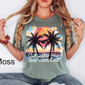 Puede incluir: Camiseta verde musgo con un dise&ntilde;o de puesta de sol, palmeras y el texto "Saltwater and sunshine". El dise&ntilde;o presenta un sol naranja y amarillo, agua azul y palmeras negras. La camiseta tiene un corte holgado.