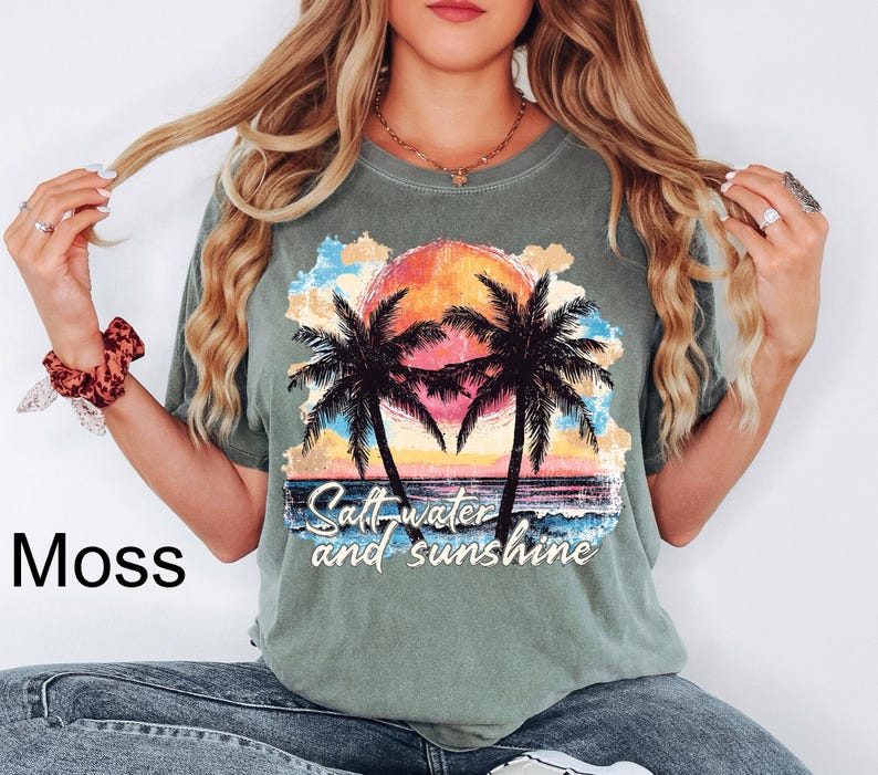 Puede incluir: Camiseta verde musgo con un dise&ntilde;o de puesta de sol, palmeras y el texto "Saltwater and sunshine". El dise&ntilde;o presenta un sol naranja y amarillo, agua azul y palmeras negras. La camiseta tiene un corte holgado.