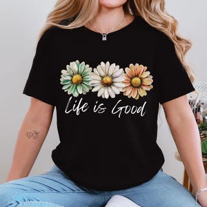 May include: Black t-shirt featuring three daisy flowers in green, white, and orange, with the cursive text "Life is Good" below the flowers. The shirt is a casual style.