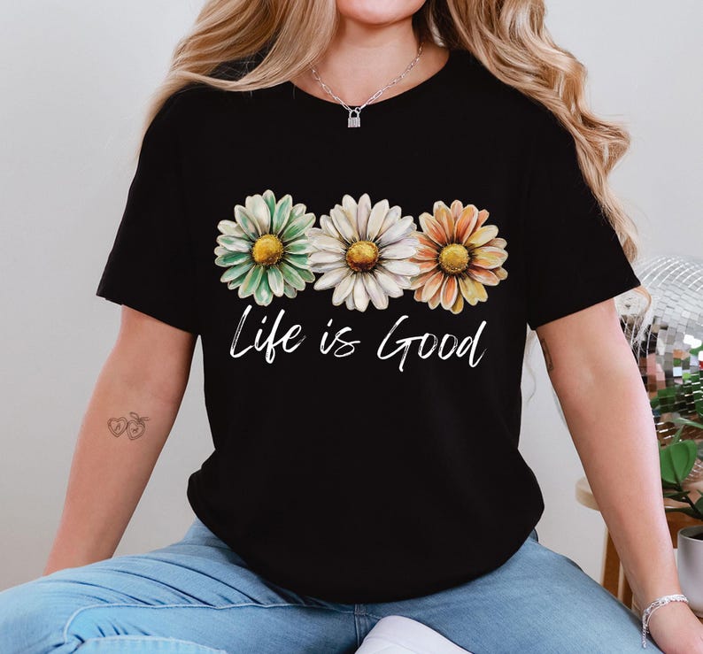 May include: Black t-shirt featuring three daisy flowers in green, white, and orange, with the cursive text "Life is Good" below the flowers. The shirt is a casual style.