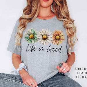 May include: Light gray t-shirt featuring three daisy flowers in green, white, and orange, with the text "Life is Good" in black script. The shirt is made of a soft, comfortable material.