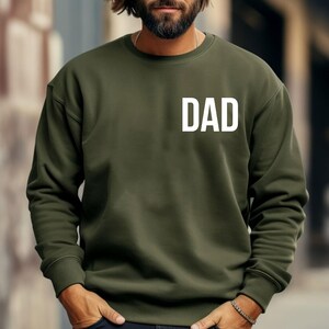 May include: Olive green sweatshirt with the white word "DAD" printed on the chest. The long-sleeved crew neck top is made of a soft material. A casual, comfortable garment.