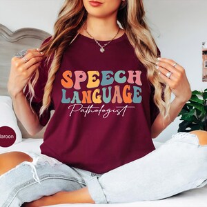 May include: Maroon t-shirt with the words "SPEECH LANGUAGE Pathologist" in a colorful, retro font. The shirt is being worn by a person sitting on a bed. The shirt is a solid maroon color.