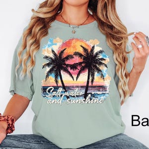 Puede incluir: Camiseta verde claro con un gr&aacute;fico colorido de una puesta de sol, palmeras y olas del oc&eacute;ano. El texto "Salt-water and sunshine" est&aacute; impreso debajo. La camiseta es de corte holgado.