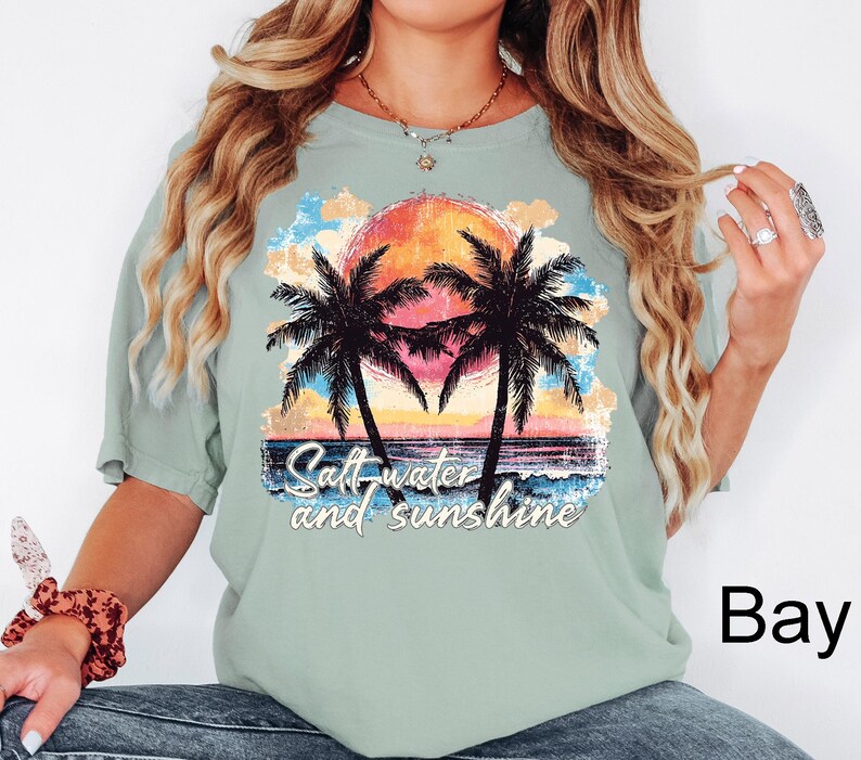Puede incluir: Camiseta verde claro con un gr&aacute;fico colorido de una puesta de sol, palmeras y olas del oc&eacute;ano. El texto "Salt-water and sunshine" est&aacute; impreso debajo. La camiseta es de corte holgado.