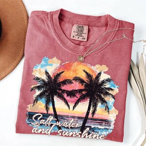 Puede incluir: Una camiseta roja con un gr&aacute;fico de una puesta de sol, palmeras y las palabras "Salt water and sunshine". La camiseta tiene una etiqueta Comfort Colors y un collar plateado. Un sombrero marr&oacute;n y una planta seca est&aacute;n en el fondo.