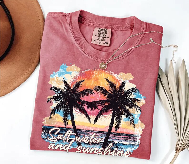 Puede incluir: Una camiseta roja con un gr&aacute;fico de una puesta de sol, palmeras y las palabras "Salt water and sunshine". La camiseta tiene una etiqueta Comfort Colors y un collar plateado. Un sombrero marr&oacute;n y una planta seca est&aacute;n en el fondo.
