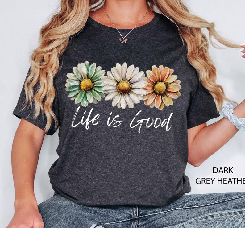 May include: Dark grey heather t-shirt featuring three daisy flowers in green, white, and orange with yellow centers. The words "Life is Good" are printed in white script below the flowers. The shirt is a casual, everyday wear item.