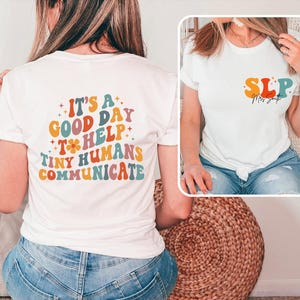 May include: White t-shirt with the text "IT'S A GOOD DAY TO HELP TINY HUMANS COMMUNICATE" in colorful retro font on the back. The front of the shirt has the letters "SLP" in a similar style. The shirt is worn with blue jeans.