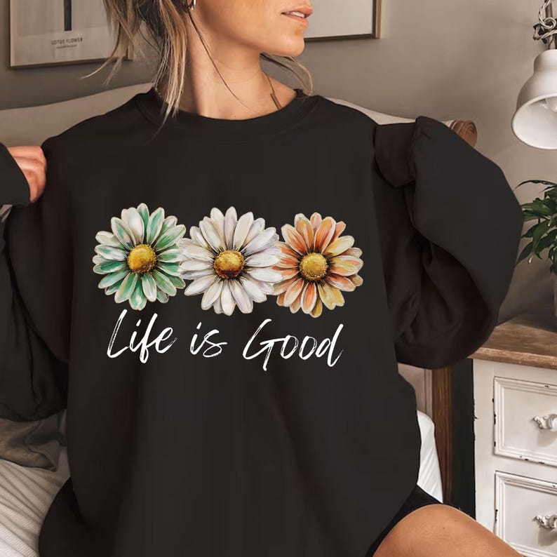 May include: Black sweatshirt featuring three daisy flowers in green, white, and orange with yellow centers. The words "Life is Good" are written in white script below the flowers. The sweatshirt is a casual style.