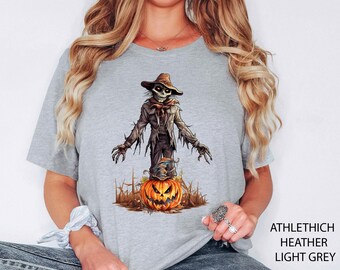 May include: Light grey t-shirt featuring a Halloween-themed graphic. The design includes a scarecrow figure emerging from a carved pumpkin. The scarecrow wears a hat and tattered clothing. The text "ATHLETHICH HEATHER LIGHT GREY" is printed on the right side of the shirt.