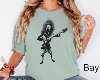 May include: A sage green short-sleeve t-shirt featuring a black graphic print of a long-haired dog with shaggy fur wearing sunglasses and playing an electric guitar. The shirt has a crew neckline and a relaxed fit, showcasing a detailed illustration.