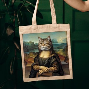 Puede incluir: Una bolsa de tela con una impresión de un gato vestido como la Mona Lisa. El gato es marrón y atigrado con ojos verdes. El fondo es una pintura de paisaje con un río y montañas.