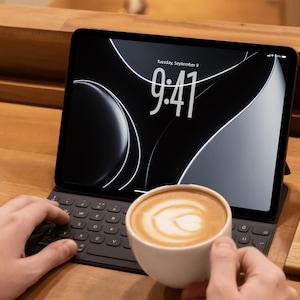 May include: A black tablet with a keyboard case displays the time 9:41 on a black and white background. A white cup of coffee with latte art is held in front of the tablet on a wooden surface.